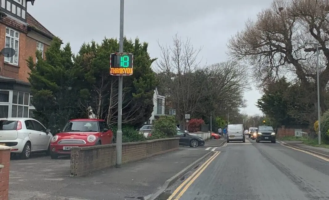 A Speed Indicator Device (SID) displaying "18" and "THANK YOU!" installed on a residential street in Burnham-On-Sea, UK, with cars parked on the side and driving on the road.